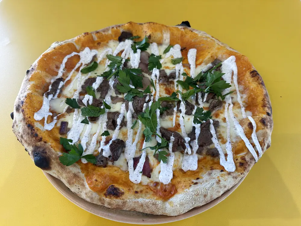 Pizza La Bosphore — Crème maison au sumac, Mozzarella fior di latte, Fines lamelles de bœuf grillé, Oignons rouges, Persil plat & menthe fraîche, Sauce au yaourt turc épicée aux herbes et à la coriandre, Huile d’olive